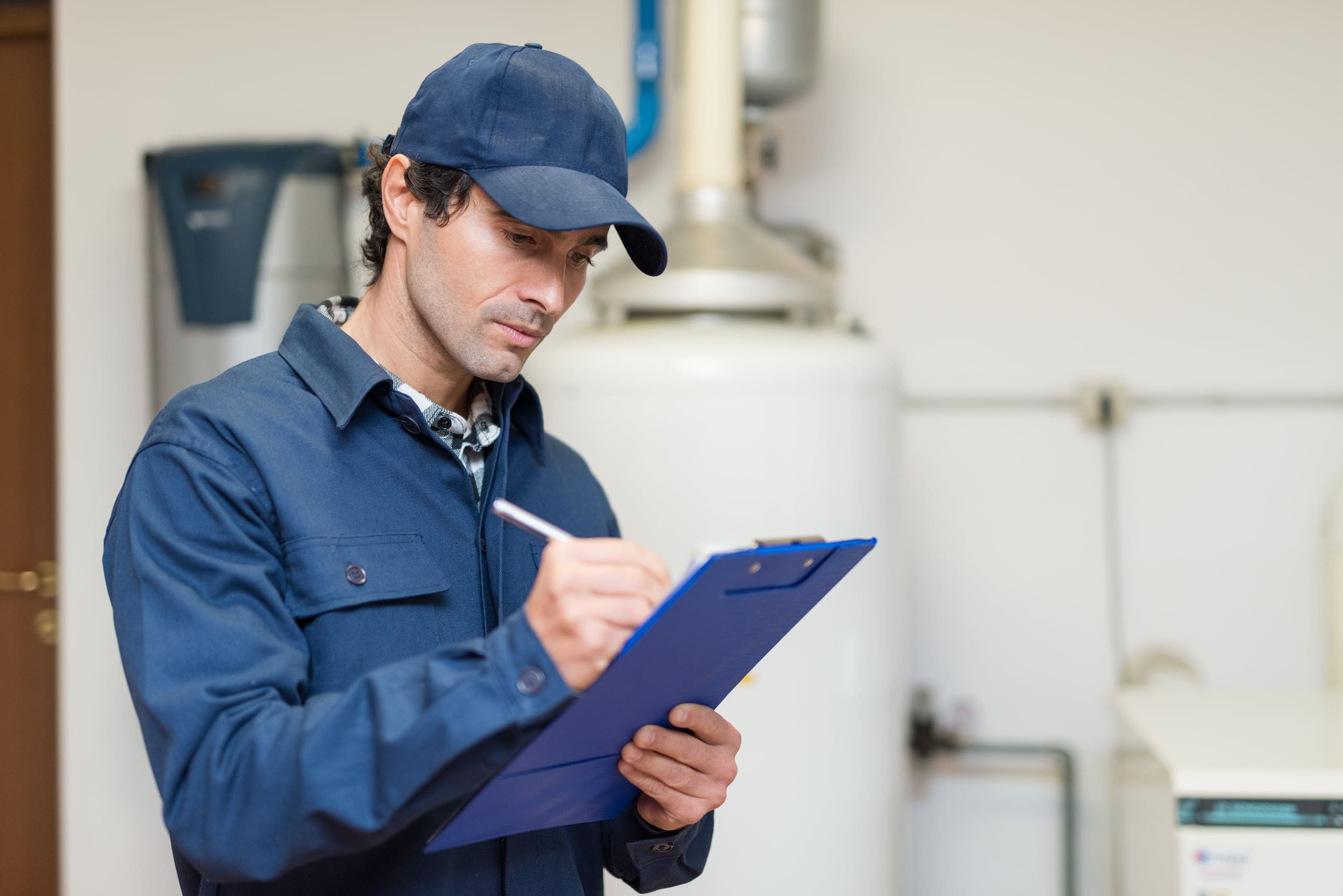 Person in a blue uniform and cap writing on a clipboard in an industrial setting with machinery in the background.