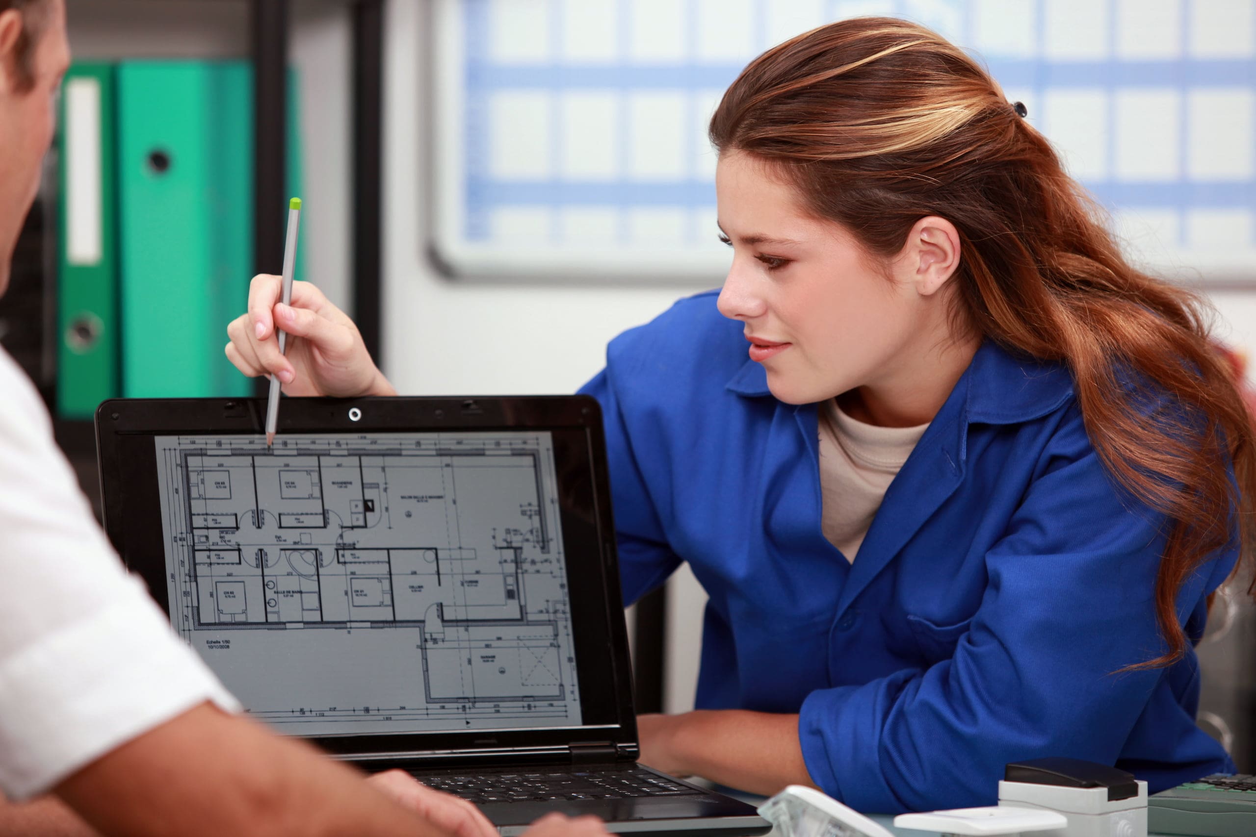 Person in a blue shirt discusses architectural plans displayed on a laptop screen with another individual.
