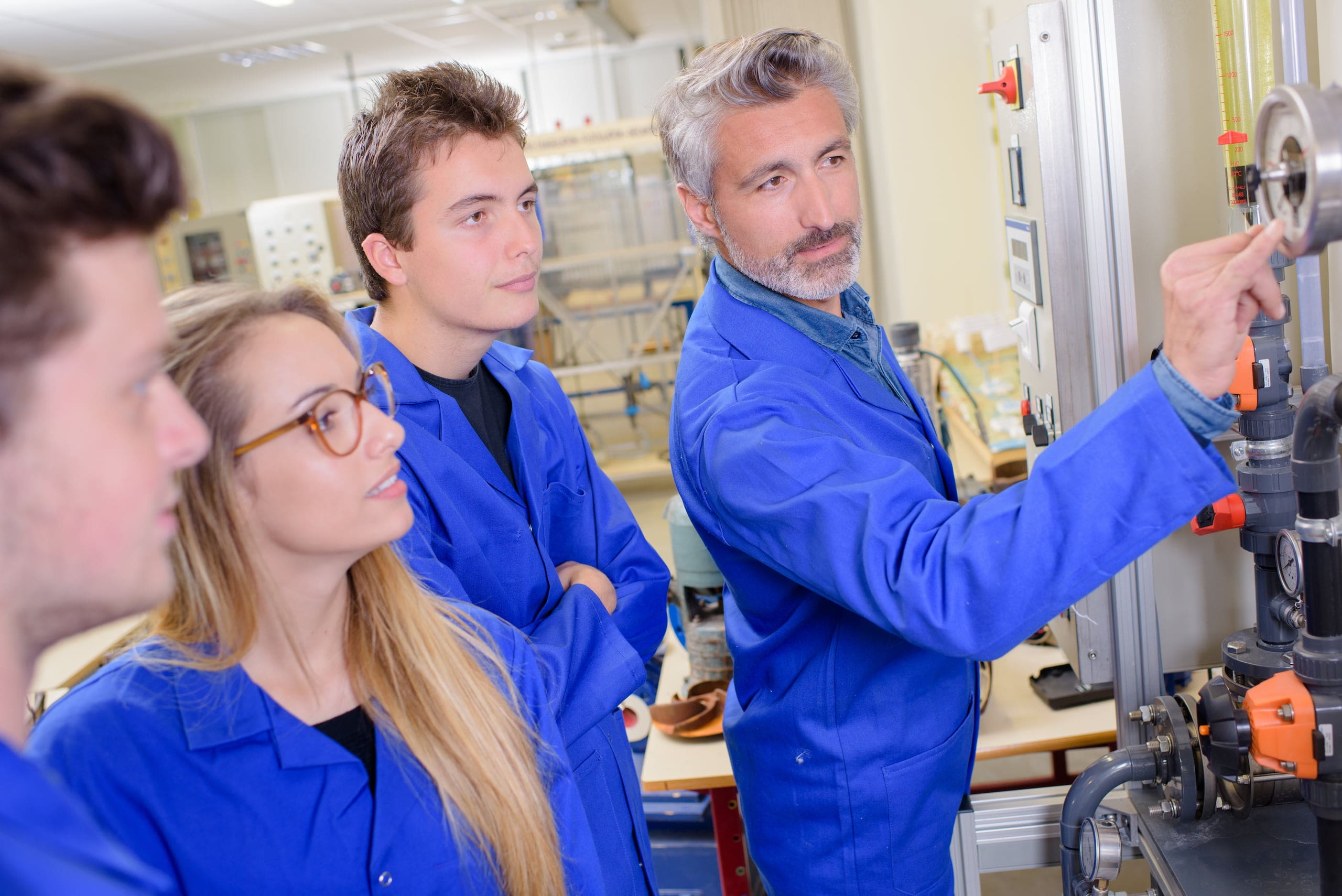 A group of four people in blue lab coats stand in a laboratory, resembling a craft teacher with students. The older man points to gauges on a machine while the others observe intently.