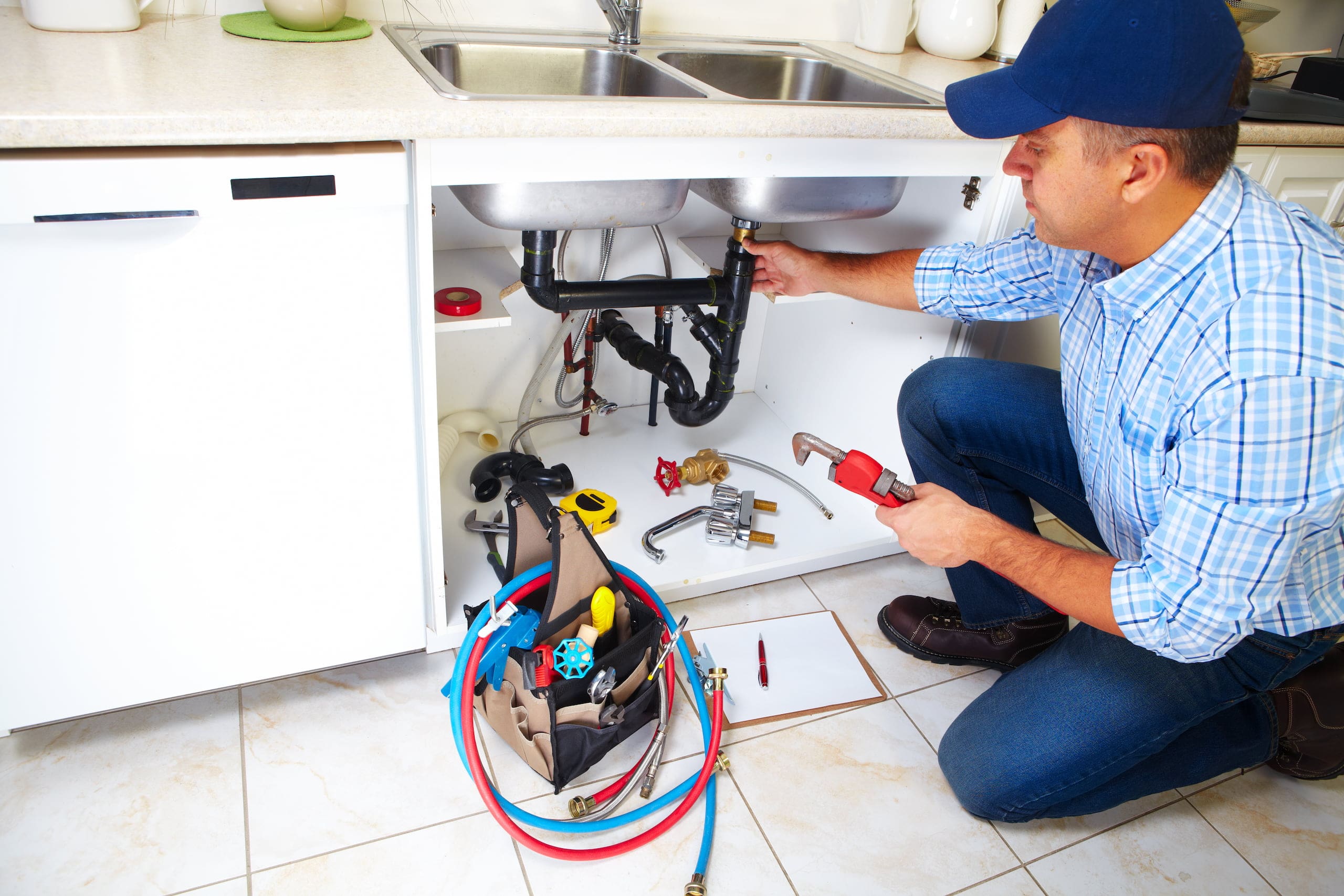 A plumber in a cap meticulously fixes the kitchen sink, tools and equipment spread on the floor beside an open tool bag.