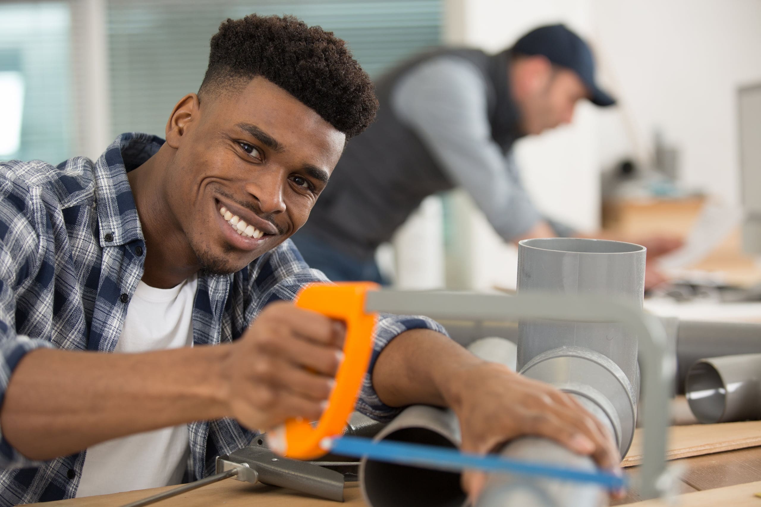 Man in a checkered shirt expertly uses a hacksaw to cut a pipe, smiling confidently at the camera. His precision rivals that of a grinder. Meanwhile, another person busies themselves at a desk in the background.