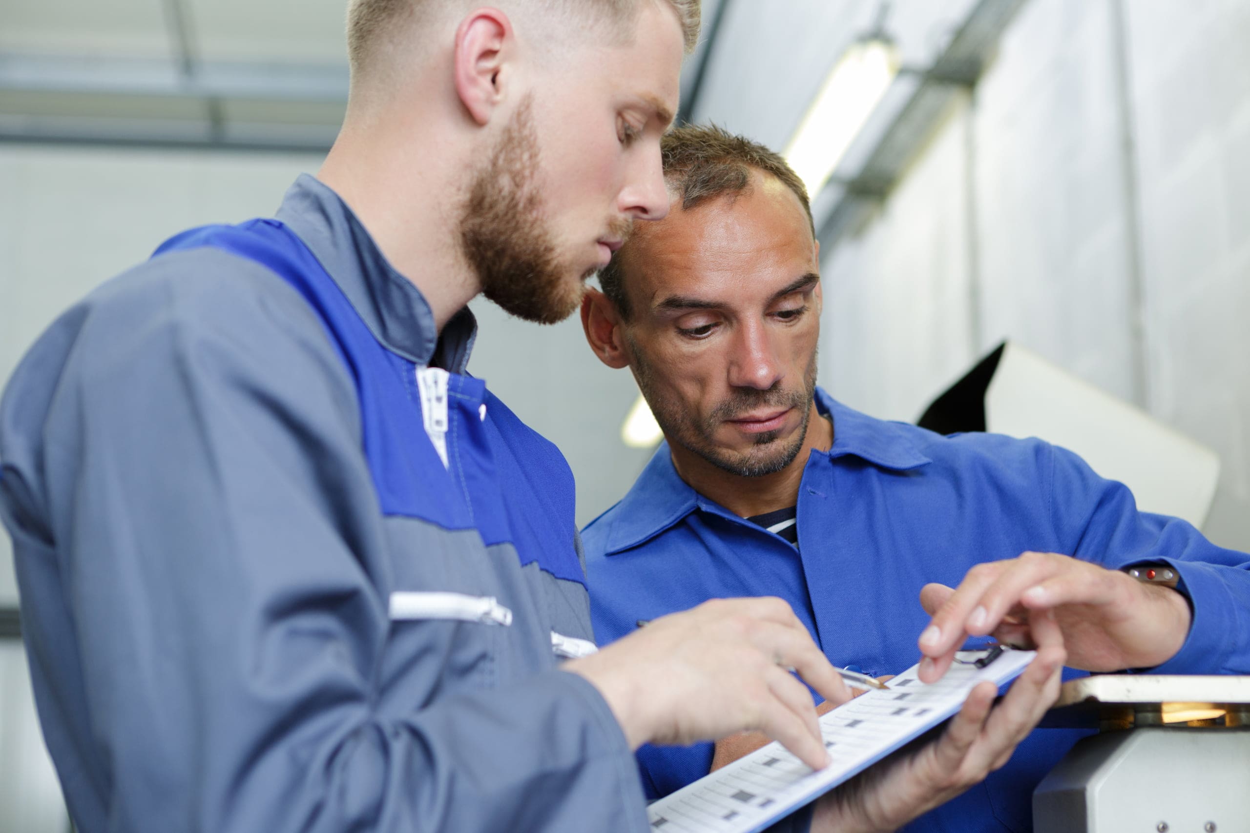 Two men in plaid shirts are examining plumbing fixtures in a white tiled room. The plumber holds a valve, explaining its role in fitting central heating, while the apprentice listens attentively.