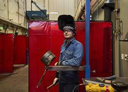 A person wearing a welding helmet and blue protective clothing stands in a workshop holding a welding tool. Red protective screens and various equipment are visible in the background.