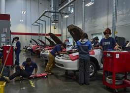 A group of mechanics working on cars in an automotive repair shop. Vehicles have their hoods open, and tools are visible on red carts.
