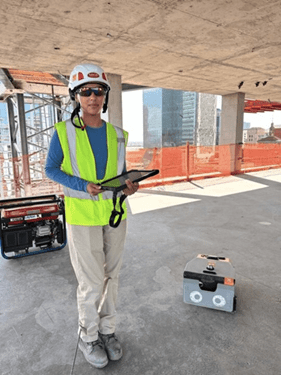 A person in a hard hat and safety vest holding a tablet on a construction site, with safety equipment and a cityscape in the background.