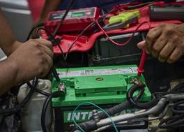 Hands holding probes on a green car battery with red and black cables attached.