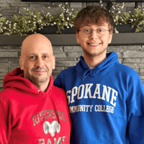 Two people stand side by side, smiling in front of a stone wall. One wears a red hoodie, the other a blue one. A decorative shelf with greenery is above them.