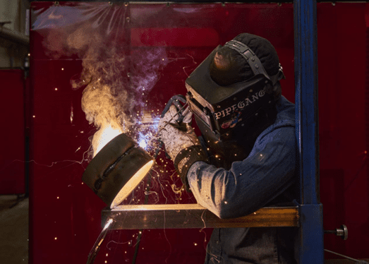 Person welding a pipe with protective gear, surrounded by sparks in an industrial setting.