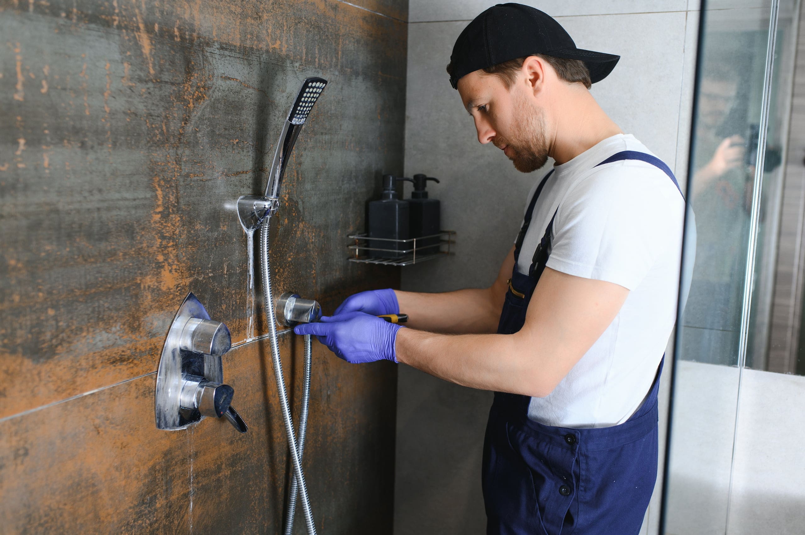 Man in overalls and gloves repairs a shower in a bathroom.