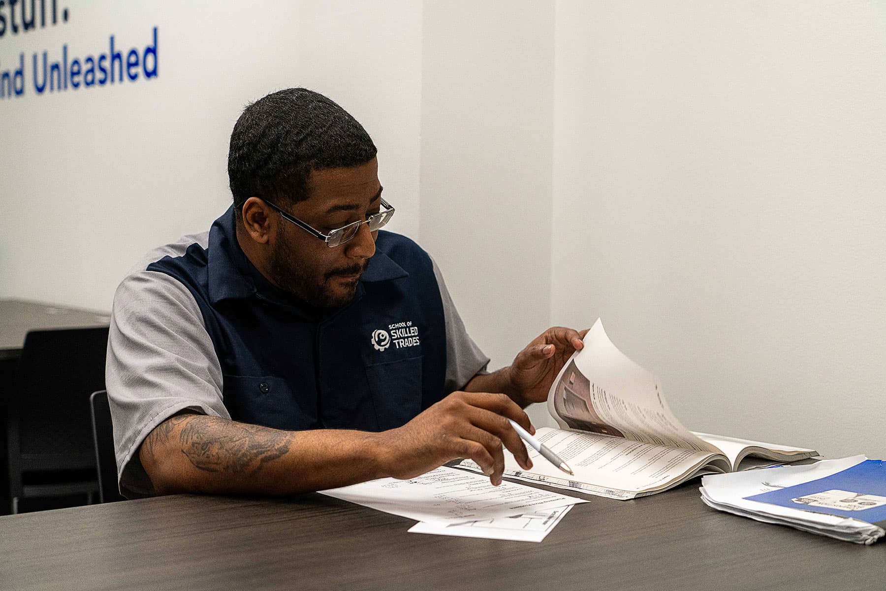 A man in work uniform sits at a table, flipping through papers and taking notes with a pen in a classroom or office setting.