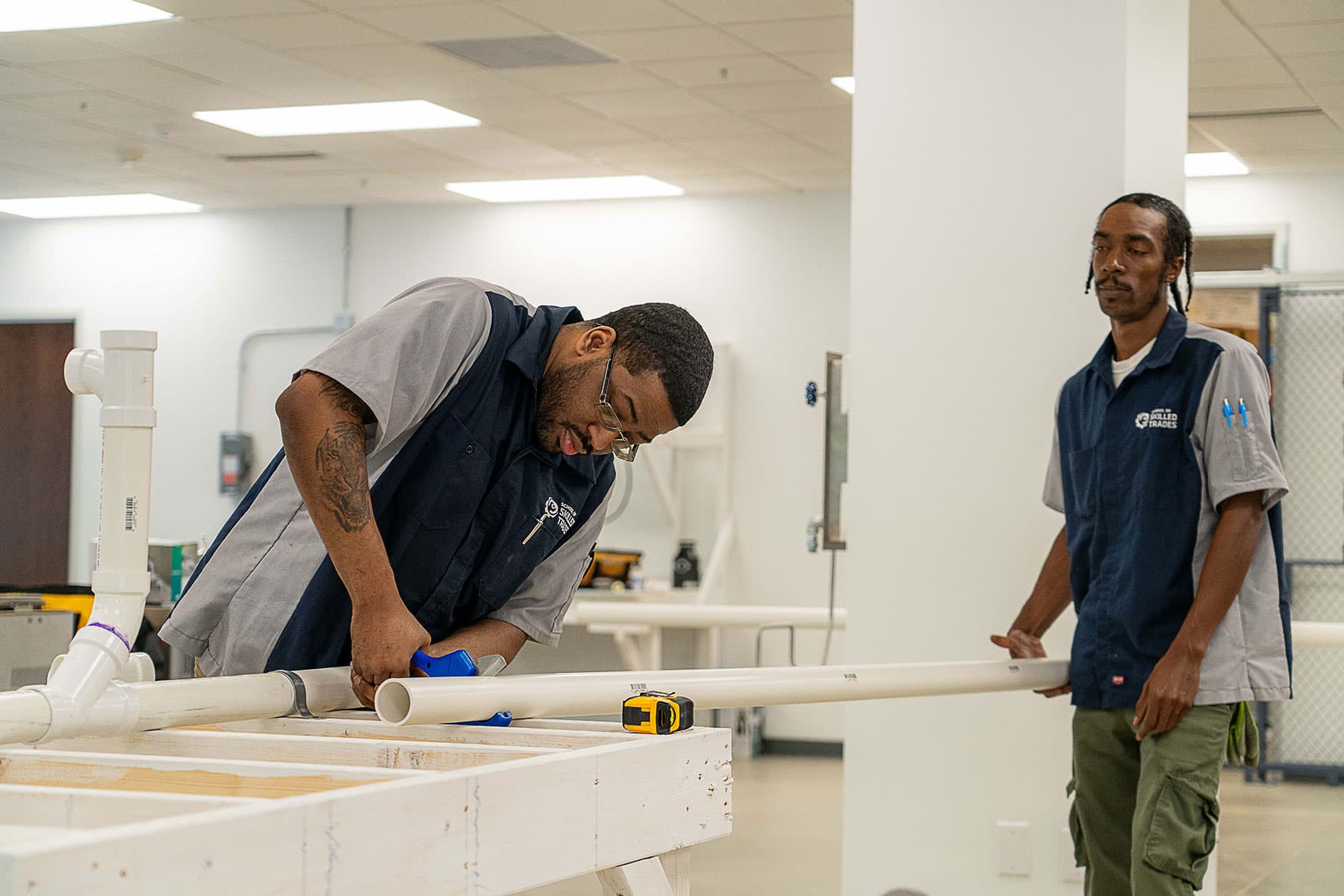 Two men in work uniforms assemble PVC pipes on a table in a workshop; one is measuring and cutting, while the other observes.
