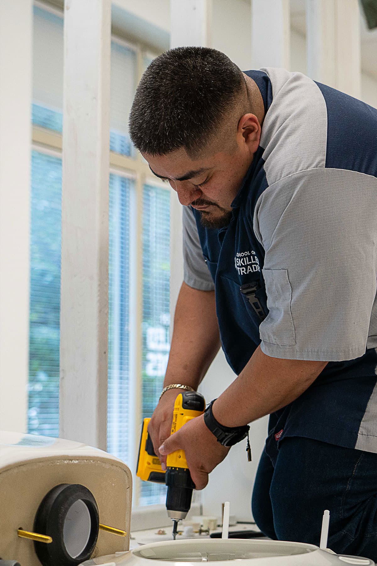 A man uses a yellow power drill to work on a white fixture inside a room with large windows.