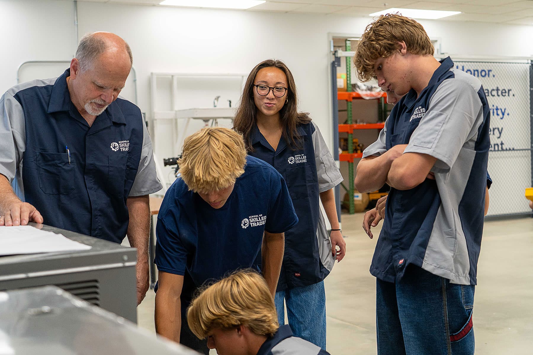 A group of students and an instructor in matching uniforms observe and work together on a machine in a classroom or workshop setting.
