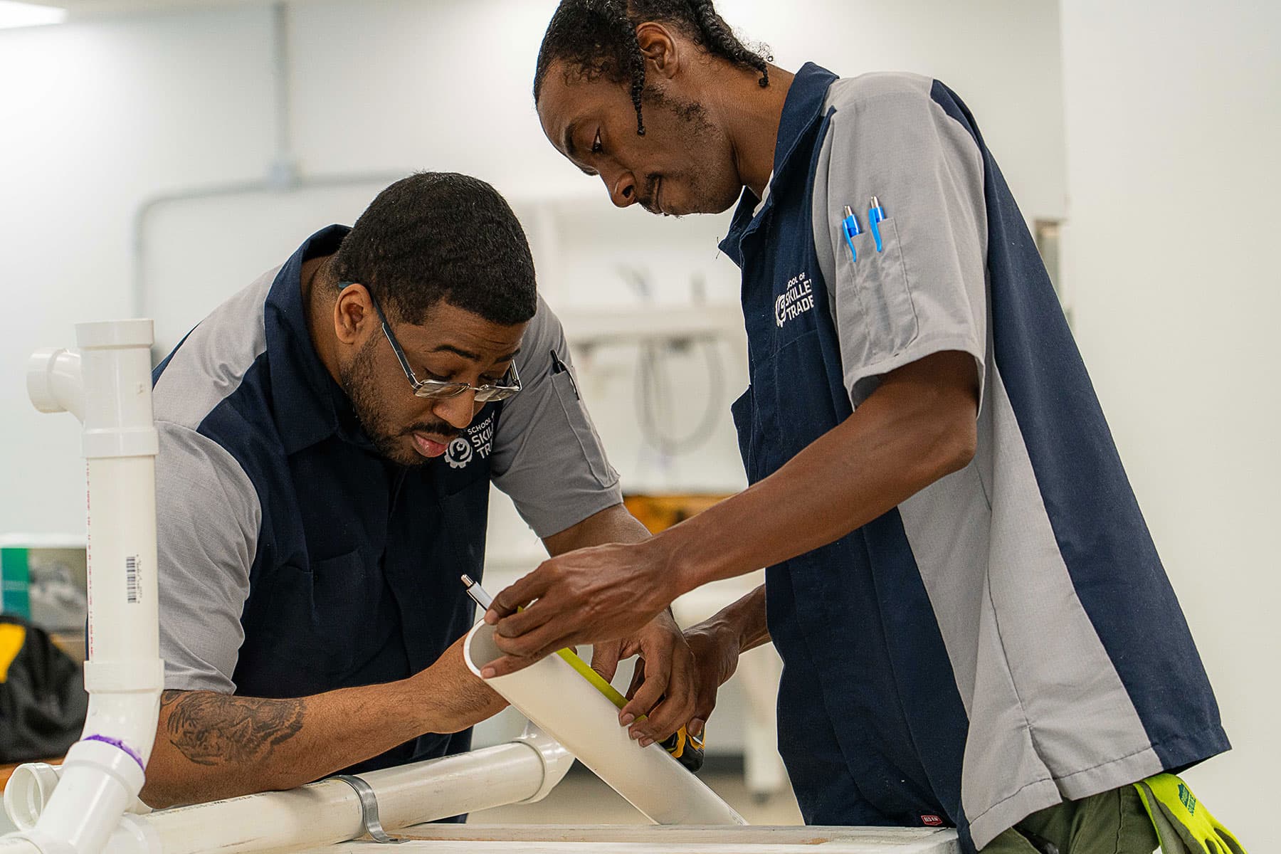 Two men in work uniforms measure and mark a white plastic pipe in a workshop setting.