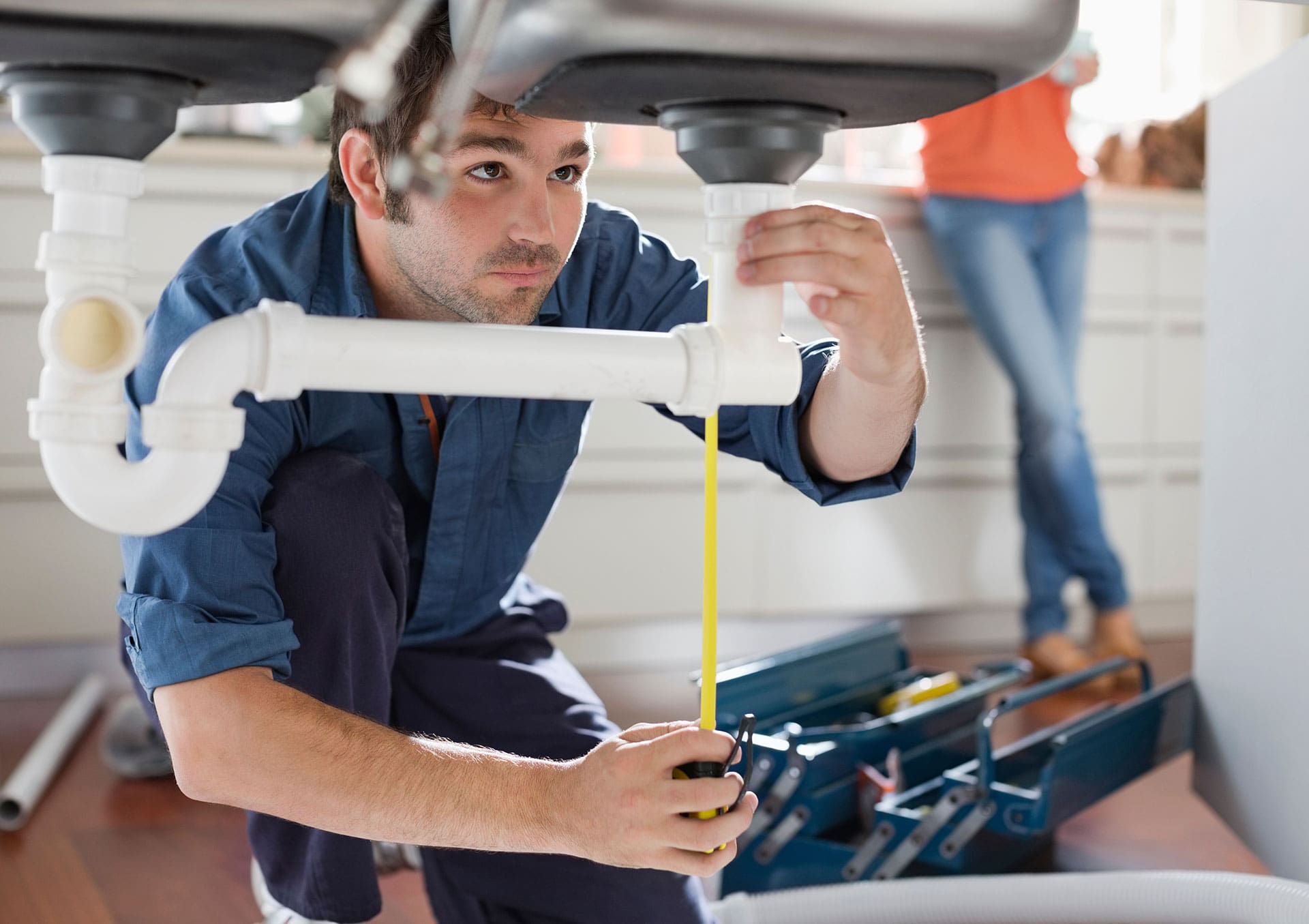A plumber kneels under a sink, inspecting the pipe with tools and an open toolbox nearby—a practical example of skills learned in trade school programs. Another person stands in the background.