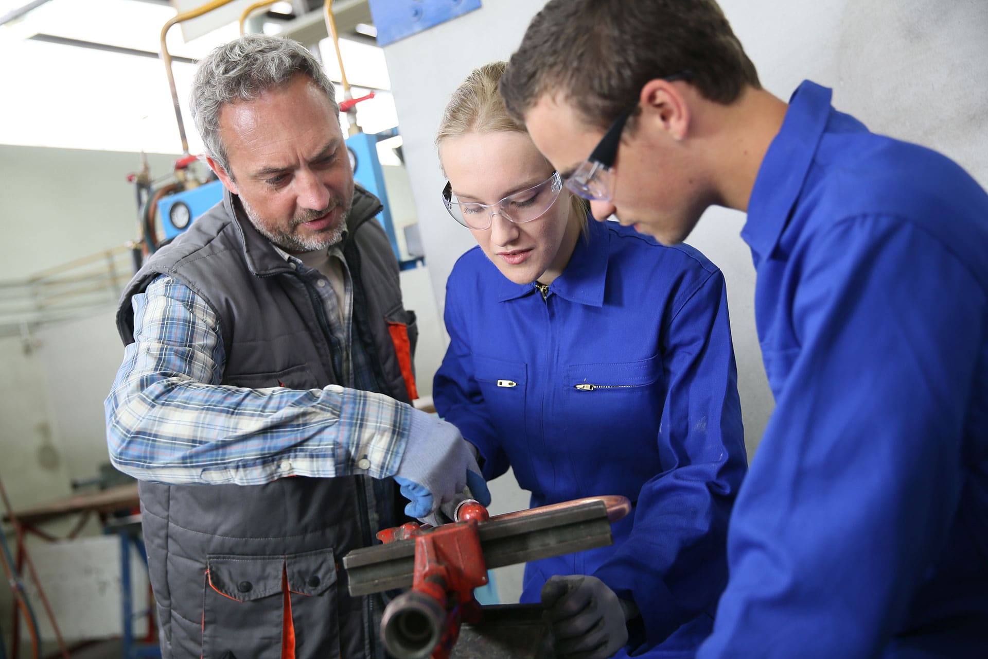 An instructor demonstrates how to use a vise to two students wearing blue coveralls and safety glasses in a workshop, showcasing hands-on training found in trade school programs.
