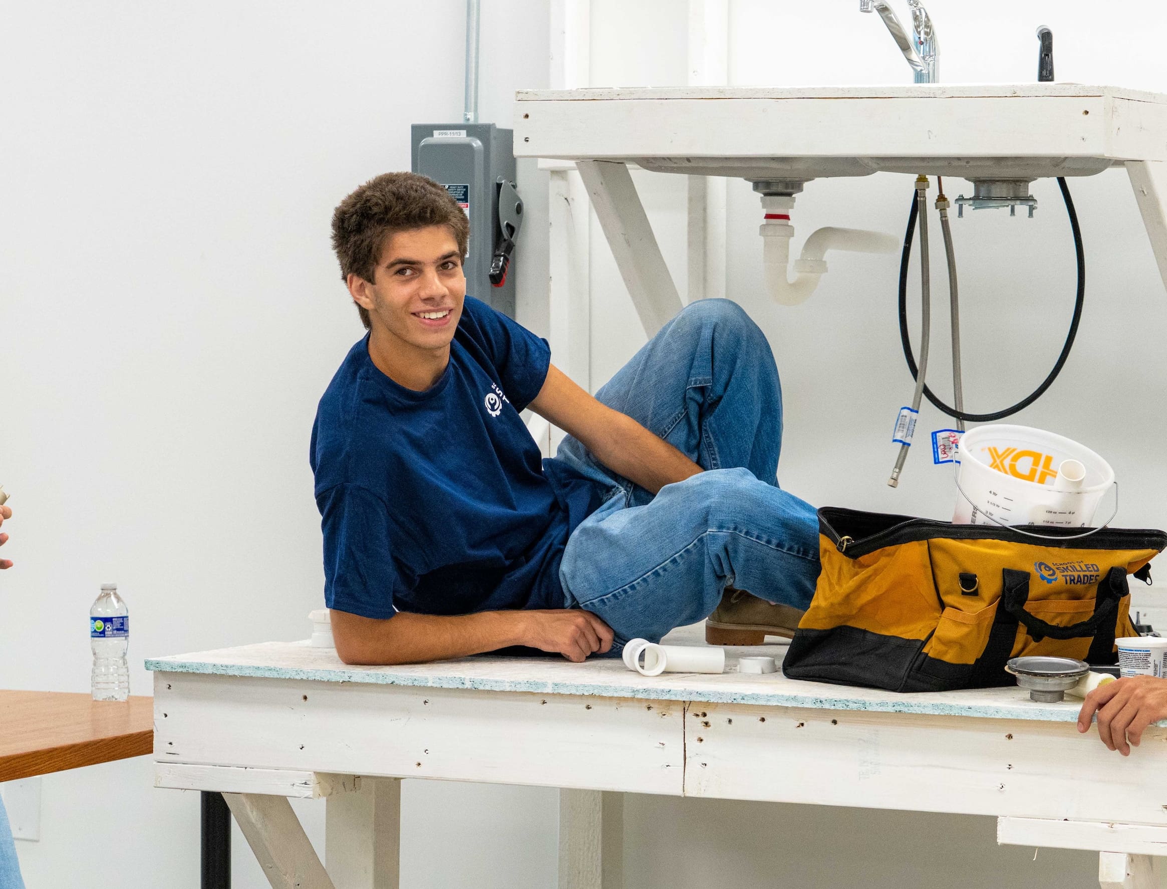A young man in a blue shirt reclines on a workbench next to plumbing tools and supplies, smiling at the camera—showcasing skills gained through trade school programs.
