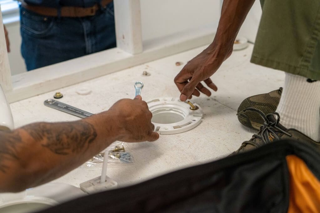 Two people are installing a toilet flange on a bathroom floor; one holds a wrench while the other adjusts a bolt, learning skills that can offset trade school cost through hands-on experience.
