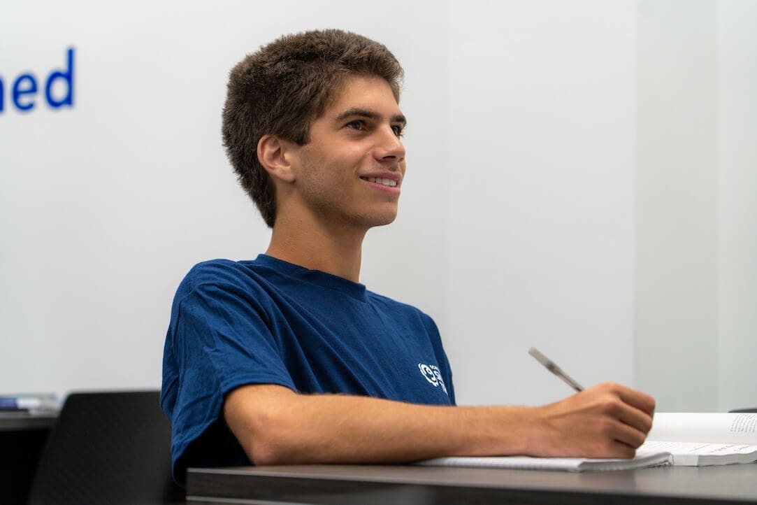 A young man in a blue T-shirt sits at a desk, smiling and holding a pen over an open notebook in a bright classroom, ready to take notes for his plumbing technician courses.