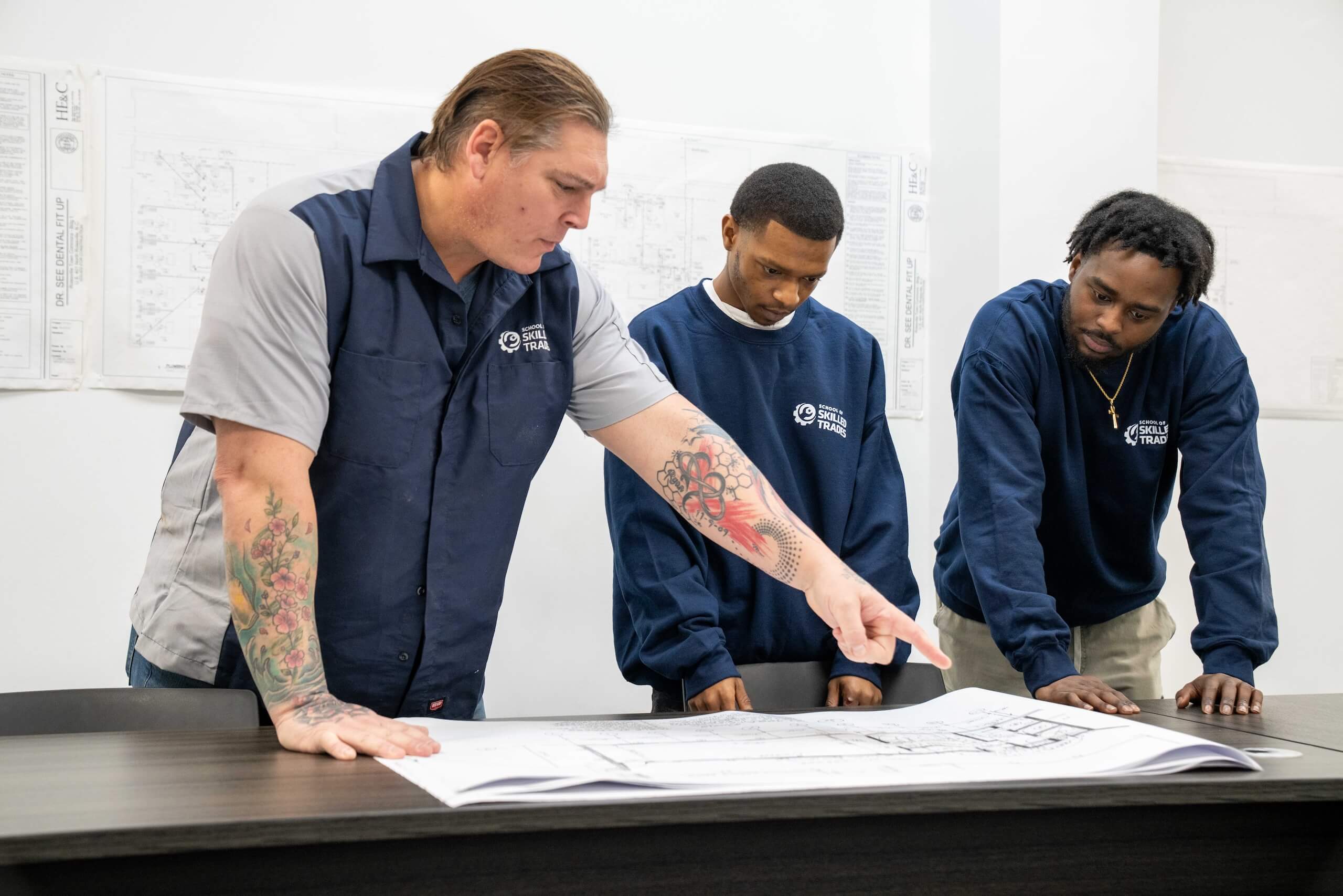 Three men stand around a table looking at blueprints; one man points at the plans while the others observe attentively.